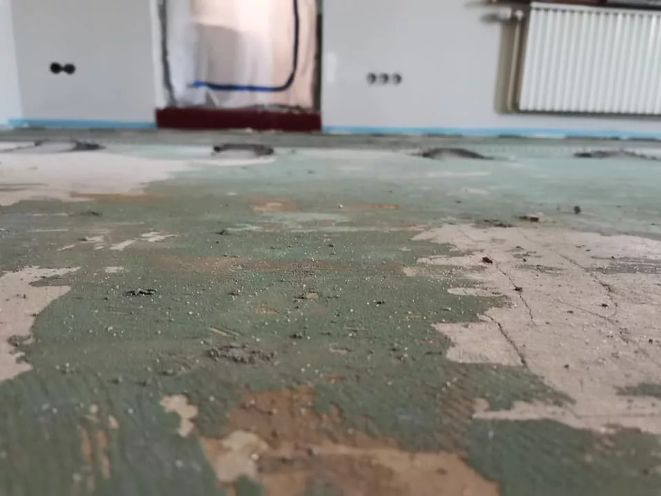 Nahaufnahme eines Bodens nach Teppichentfernung mit sichtbaren Teppichresten und Staubkörnern, im Hintergrund eine Staubschutzfolie.